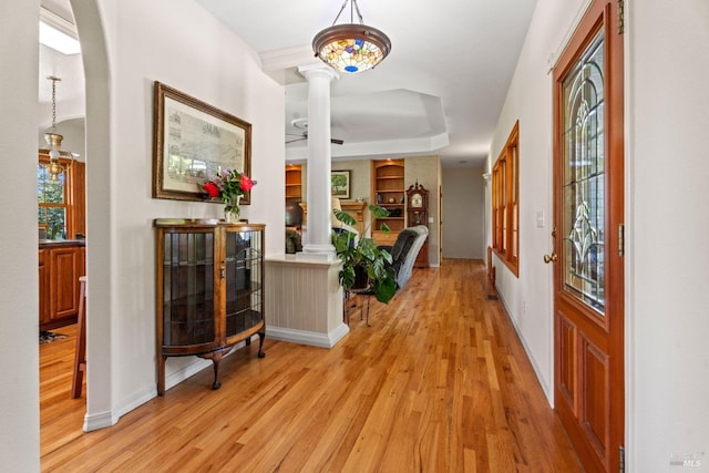 hall featuring baseboards, a raised ceiling, light wood-style flooring, and ornate columns