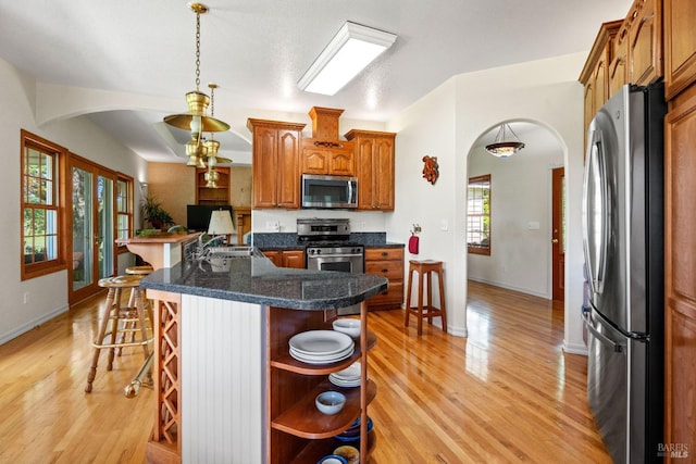 kitchen featuring brown cabinetry, light wood-type flooring, arched walkways, stainless steel appliances, and a kitchen bar