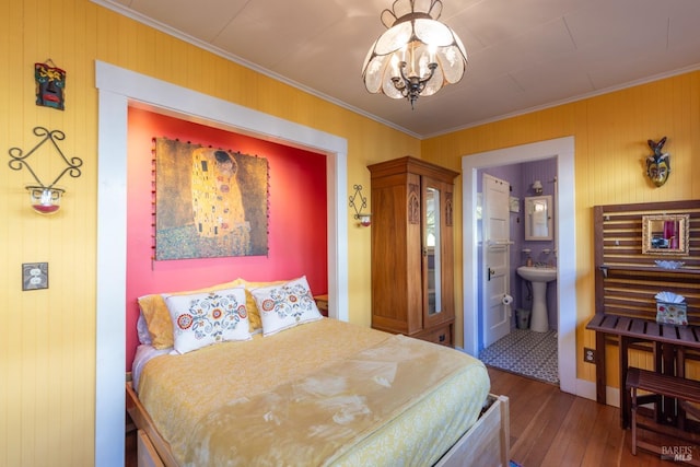 bedroom with hardwood / wood-style floors, wood walls, an inviting chandelier, connected bathroom, and crown molding