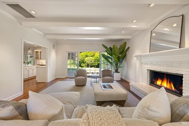 living area featuring crown molding, wood finished floors, baseboards, a brick fireplace, and beam ceiling