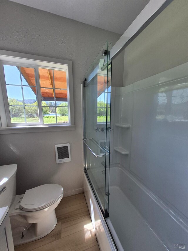 bathroom with toilet, baseboards, heating unit, wood finished floors, and combined bath / shower with glass door