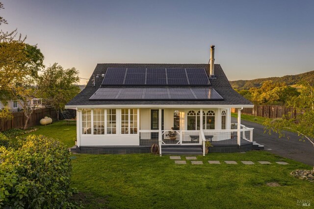 back of house at dusk featuring roof mounted solar panels, a lawn, a porch, and fence