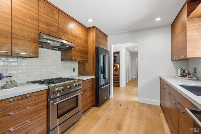 kitchen featuring brown cabinetry, premium appliances, under cabinet range hood, and tasteful backsplash