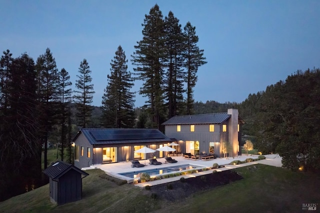back of house with board and batten siding, a chimney, an outdoor pool, roof mounted solar panels, and a patio