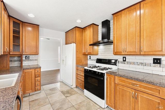 kitchen with white refrigerator with ice dispenser, decorative backsplash, wall chimney range hood, brown cabinets, and range with gas cooktop