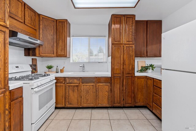 kitchen featuring light tile patterned floors, brown cabinets, a sink, under cabinet range hood, and white appliances