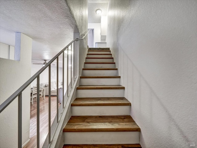 staircase featuring a textured ceiling, a textured wall, wood finished floors, and a ceiling fan