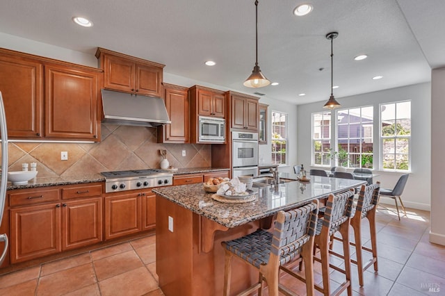 kitchen featuring under cabinet range hood, stainless steel appliances, brown cabinetry, and dark stone countertops