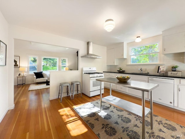 kitchen featuring wall chimney range hood, dark countertops, a sink, white cabinets, and white appliances