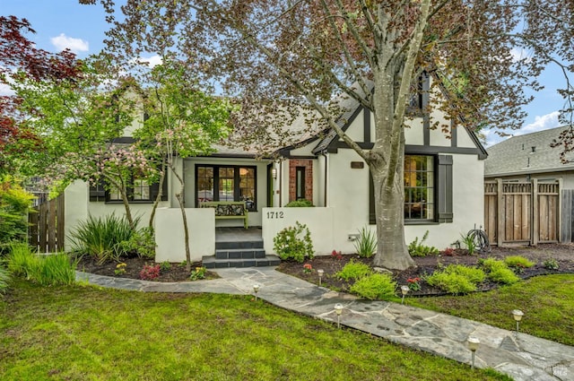 tudor home featuring a porch, a front yard, stucco siding, and fence