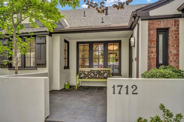 doorway to property featuring brick siding, a shingled roof, and stucco siding