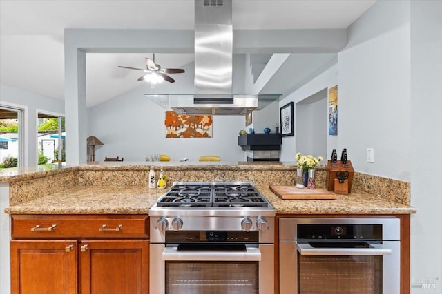 kitchen with a ceiling fan, island range hood, appliances with stainless steel finishes, brown cabinets, and light stone counters