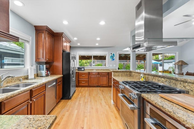 kitchen featuring light stone countertops, island exhaust hood, a sink, light wood-style flooring, and appliances with stainless steel finishes