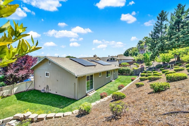 back of house with a fenced backyard, roof with shingles, a yard, and solar panels
