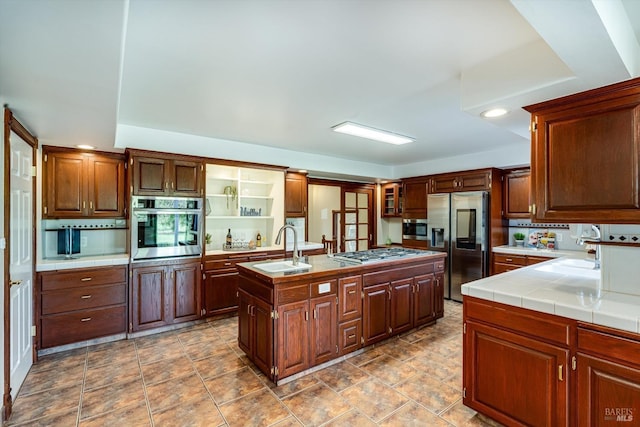 kitchen with a sink, open shelves, a center island with sink, tile countertops, and appliances with stainless steel finishes