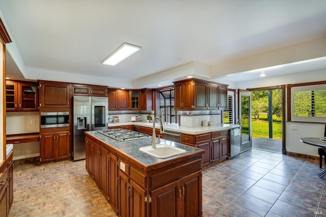 kitchen with glass insert cabinets, appliances with stainless steel finishes, a sink, baseboards, and a center island with sink