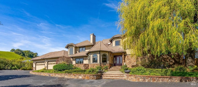 view of front facade featuring a tile roof, a garage, a chimney, aphalt driveway, and stucco siding