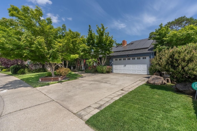 view of property hidden behind natural elements with an attached garage, a front lawn, a chimney, solar panels, and driveway