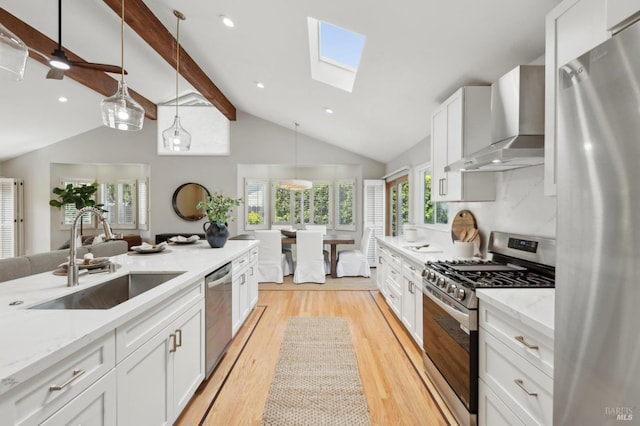 kitchen with wall chimney range hood, appliances with stainless steel finishes, a sink, plenty of natural light, and lofted ceiling with skylight