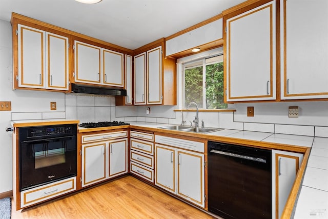 kitchen with tile counters, light wood-type flooring, under cabinet range hood, a sink, and black appliances