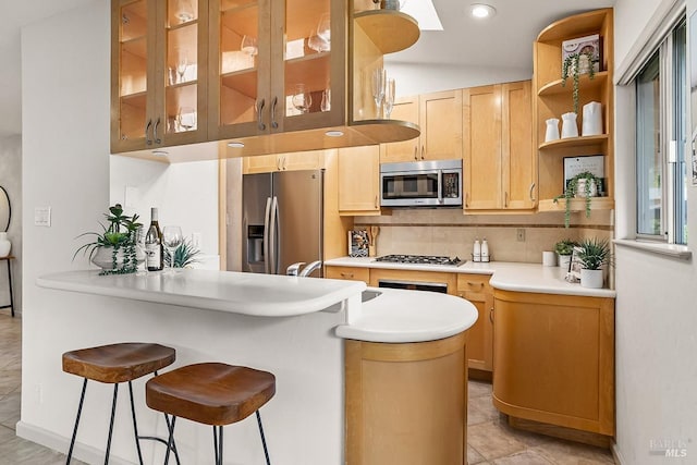 kitchen featuring light brown cabinetry, glass insert cabinets, a kitchen breakfast bar, a sink, and appliances with stainless steel finishes