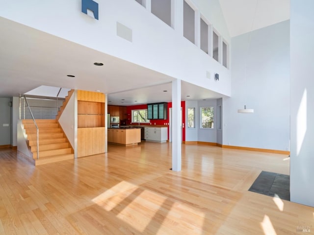 unfurnished living room with baseboards, light wood-style flooring, a towering ceiling, and stairs
