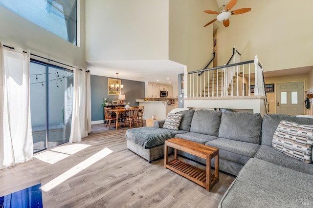 living room featuring stairs, light wood-style flooring, ceiling fan with notable chandelier, and a high ceiling
