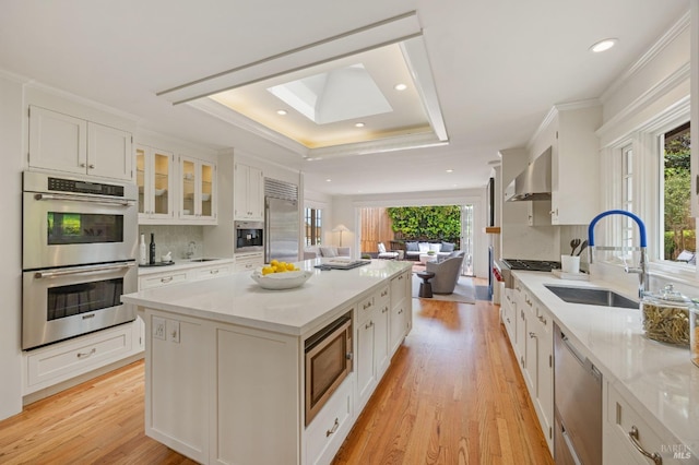 kitchen with white cabinets, a sink, built in appliances, wall chimney exhaust hood, and a tray ceiling