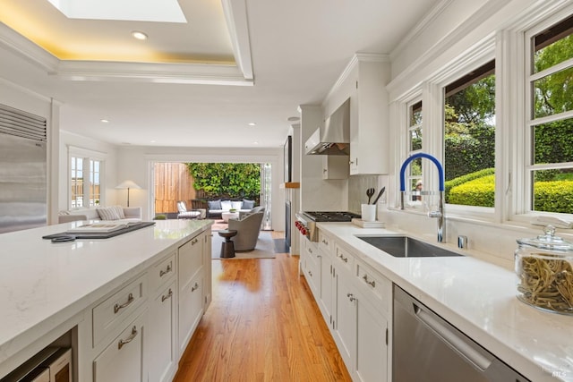 kitchen featuring appliances with stainless steel finishes, a sink, wall chimney exhaust hood, white cabinetry, and light wood-type flooring