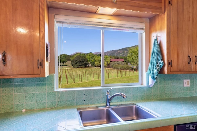 kitchen featuring brown cabinets, a sink, tile countertops, decorative backsplash, and a mountain view