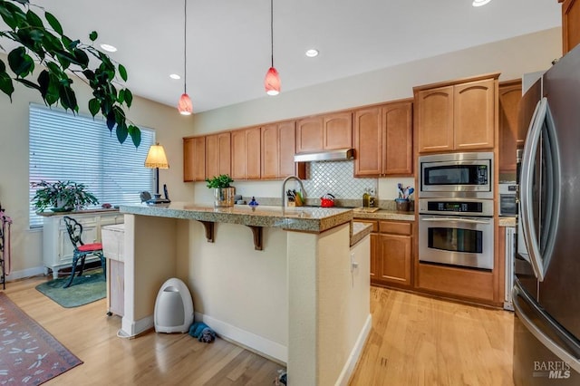 kitchen featuring a kitchen island with sink, light wood-style flooring, stainless steel appliances, decorative backsplash, and under cabinet range hood