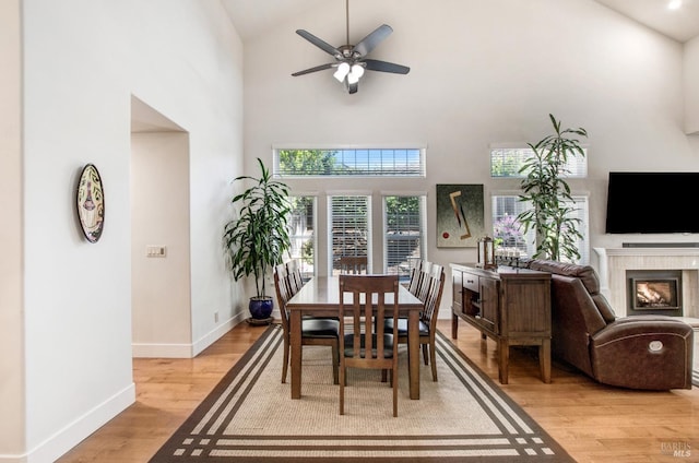dining room with ceiling fan, high vaulted ceiling, a glass covered fireplace, light wood-style flooring, and baseboards