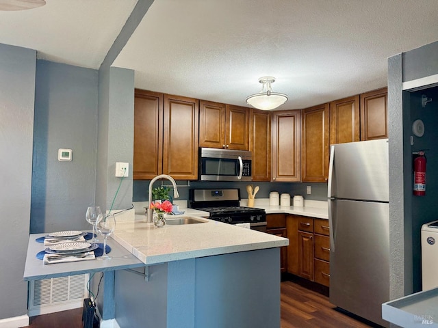 kitchen with brown cabinetry, dark wood finished floors, a sink, a peninsula, and stainless steel appliances