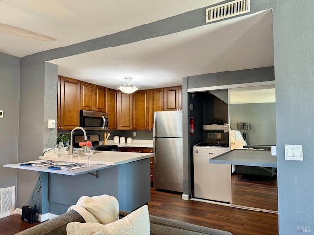 kitchen with brown cabinetry, visible vents, a peninsula, dark wood finished floors, and appliances with stainless steel finishes