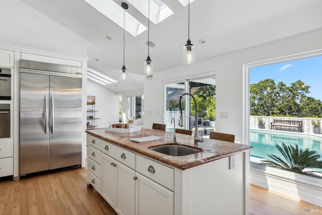 kitchen featuring vaulted ceiling with skylight, stainless steel built in fridge, hanging light fixtures, dark stone countertops, and light wood-style flooring