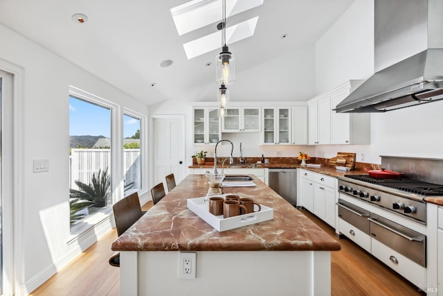 kitchen with wall chimney range hood, white cabinets, appliances with stainless steel finishes, a sink, and a skylight