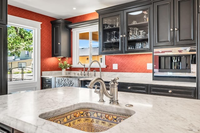 kitchen featuring dark cabinetry, a sink, light stone counters, and glass insert cabinets