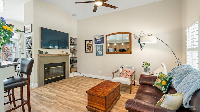 living room with a glass covered fireplace, wood finished floors, baseboards, ceiling fan, and high vaulted ceiling
