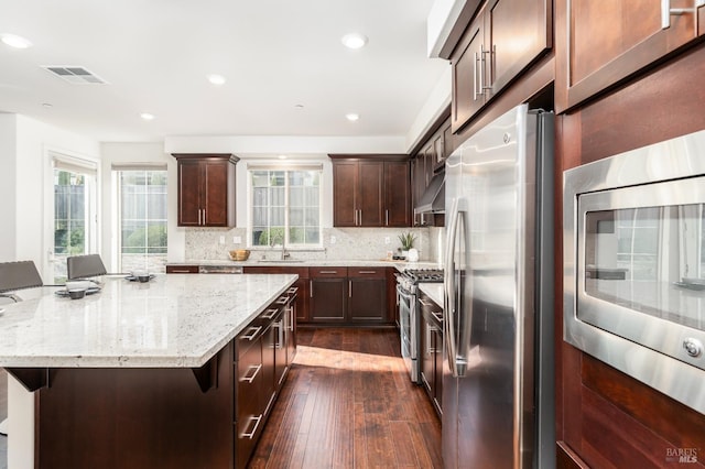kitchen featuring backsplash, dark brown cabinets, light stone counters, stainless steel appliances, and dark wood-type flooring