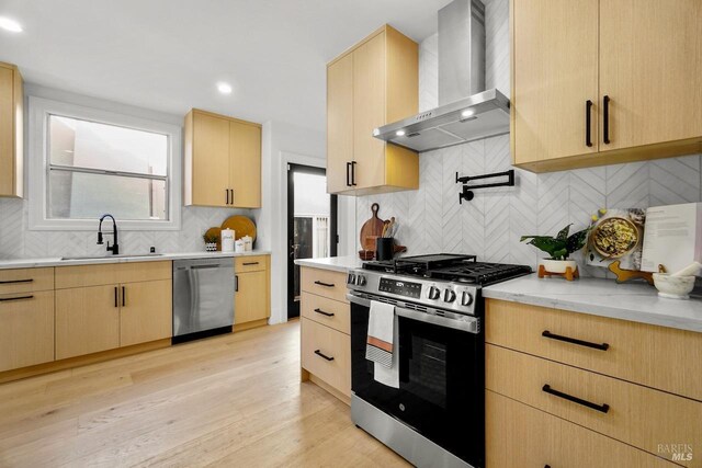 kitchen with wall chimney range hood, appliances with stainless steel finishes, a sink, and light brown cabinets