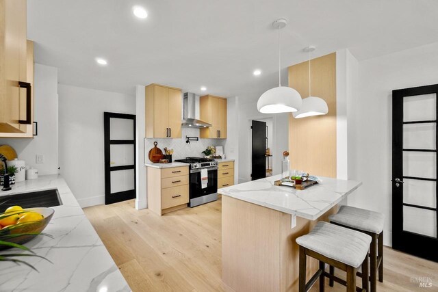 kitchen with light brown cabinetry, light wood finished floors, stainless steel range with gas stovetop, wall chimney exhaust hood, and tasteful backsplash