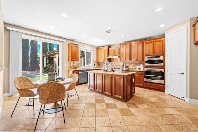 kitchen with decorative backsplash, stone tile floors, dishwasher, under cabinet range hood, and an island with sink