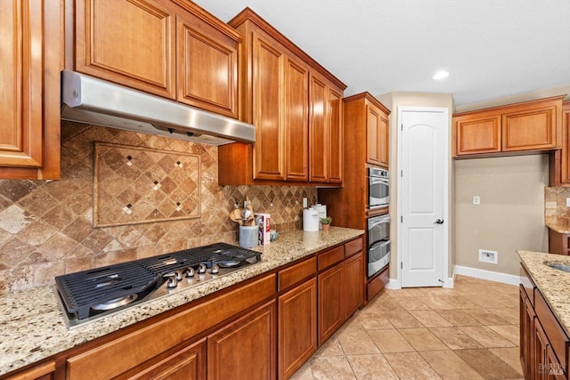 kitchen with light stone countertops, brown cabinets, baseboards, under cabinet range hood, and stainless steel appliances