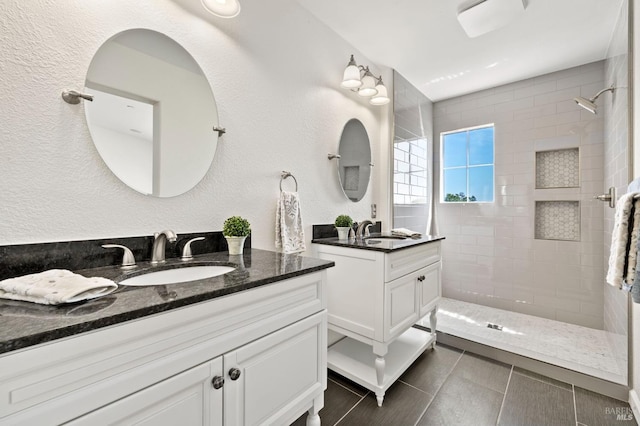 full bath featuring a textured wall, tiled shower, two vanities, and a sink