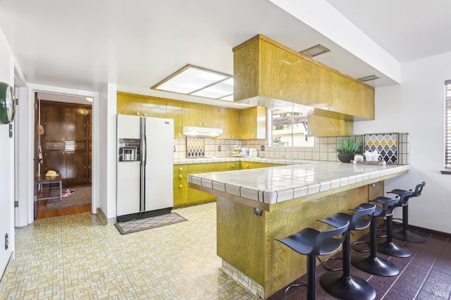 kitchen with decorative backsplash, white fridge with ice dispenser, tile counters, and a peninsula