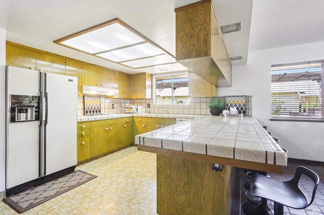 kitchen featuring decorative backsplash, a peninsula, white appliances, tile counters, and under cabinet range hood
