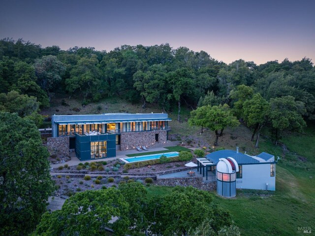 back of house at dusk featuring an outdoor pool, a patio area, and a wooded view