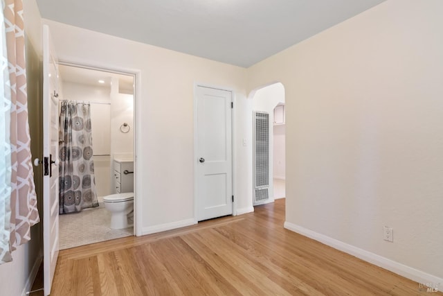 unfurnished bedroom featuring connected bathroom, light wood-style floors, arched walkways, a heating unit, and baseboards