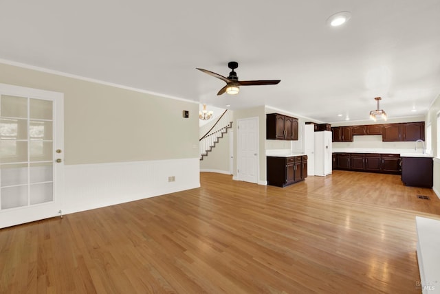 unfurnished living room featuring a ceiling fan, wainscoting, light wood-type flooring, stairway, and ornamental molding