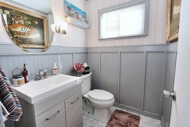 half bath with wainscoting, vanity, toilet, marble finish floor, and a decorative wall